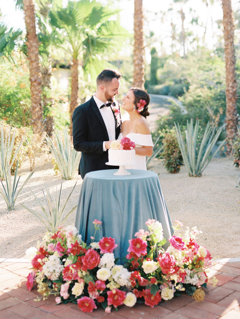 Married couple posing behind cake table with colorful flowers surrounding.