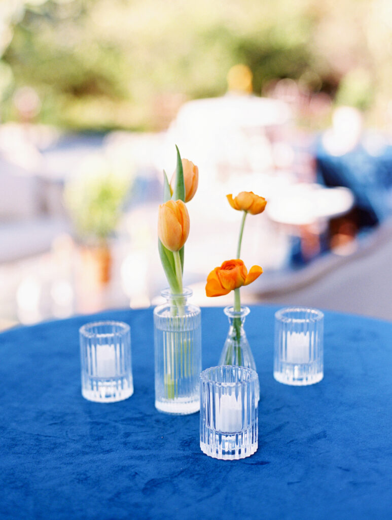 An outdoor wedding reception cocktail table with a blue velvet table cloth, glass bud vases with orange blooms, and votive candles 