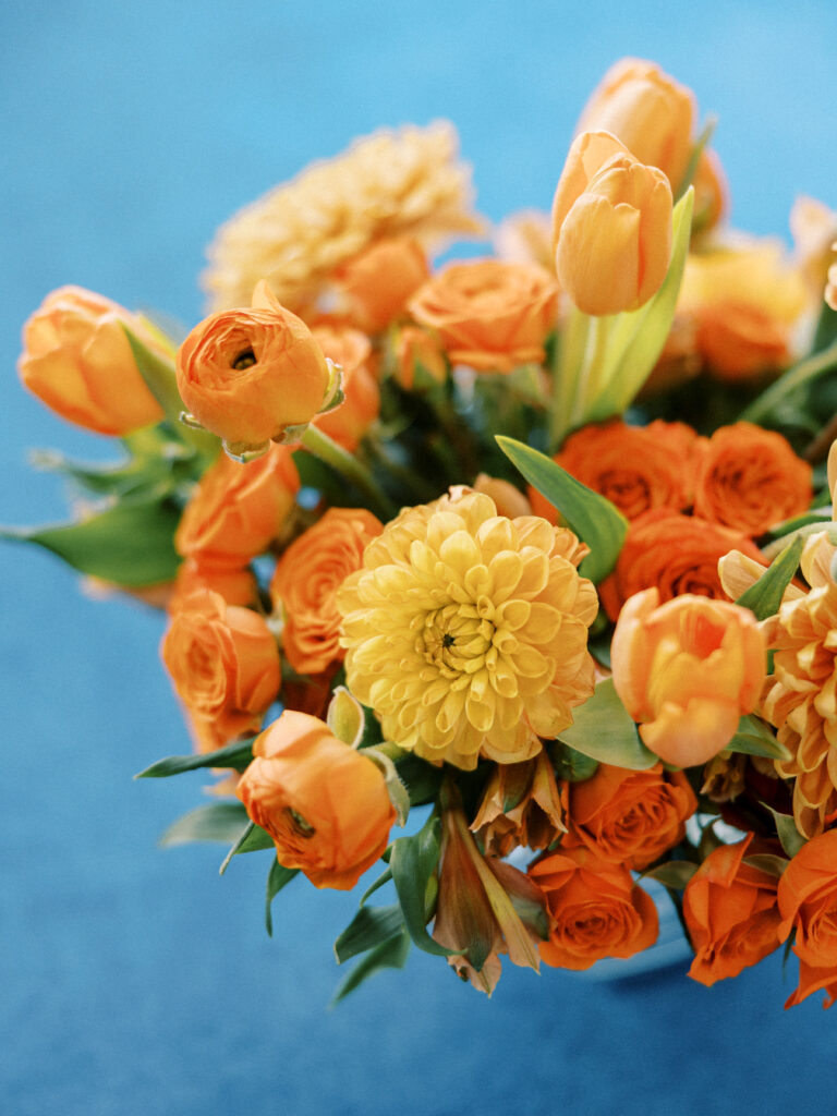 A lush floral arrangement made with vibrant orange florals, including spray roses, tulips, and Dahlias sitting on a blue velvet table cloth 