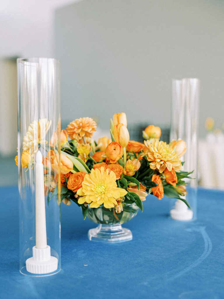 A lush floral arrangement made with vibrant orange florals, including spray roses, tulips, and Dahlias sitting next to two taper candles on a blue velvet table cloth 