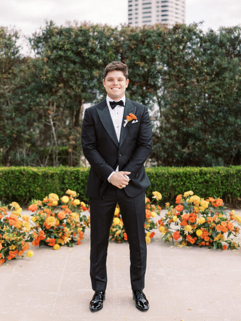 Groom standing at wedding ceremony alter surrounded by a semi circle of ground floral arrangements made with orange flowers