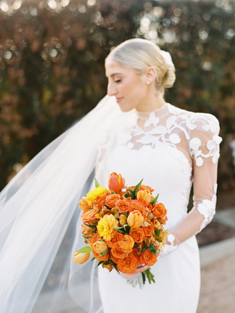 Beautiful bride with trialing veil holding a bridal bouquet made of vibrant orange flowers