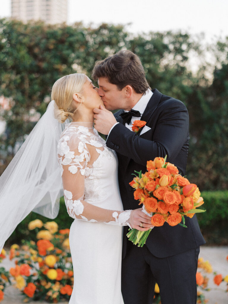 An outdoor wedding ceremony set up with bride and groom kissing at the alter surrounded by a semi circle of orange flower arrangements