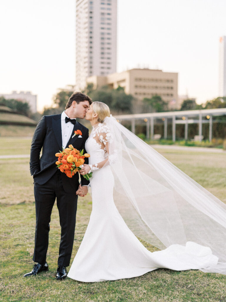 Bride and groom kissing while holding a colorful bridal bouquet made with orange flowers