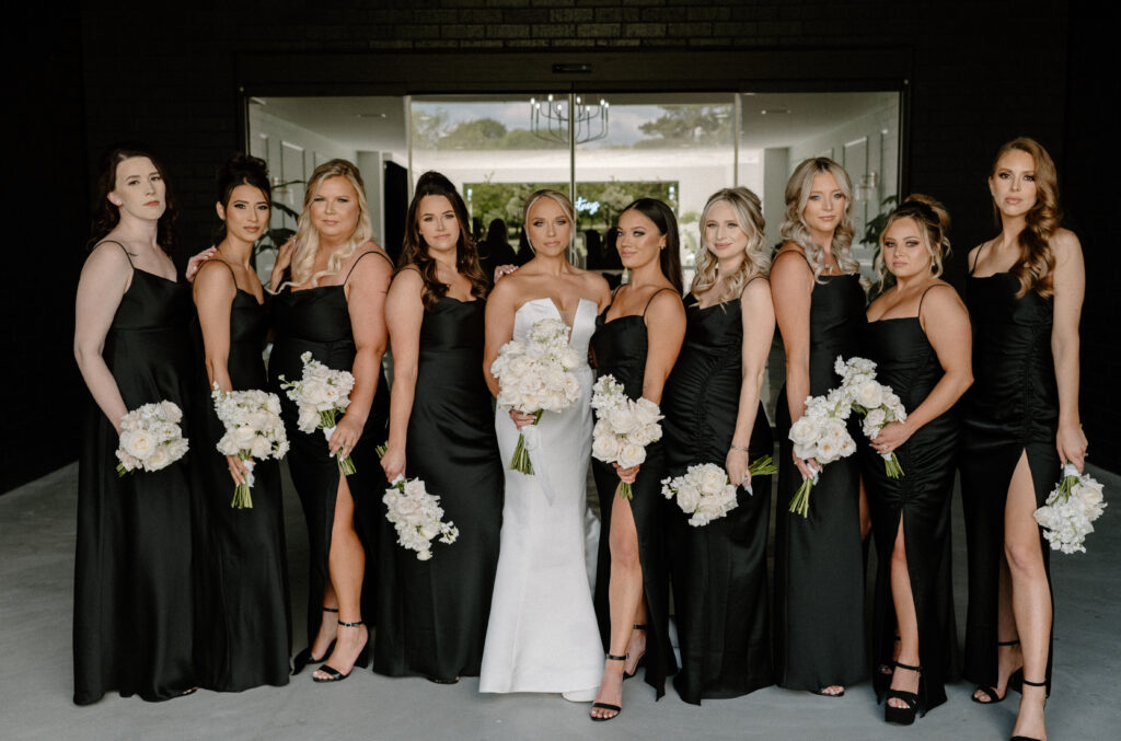 Bride posing with her bridesmaids wearing all black dresses and holding their bouquets made with all white roses