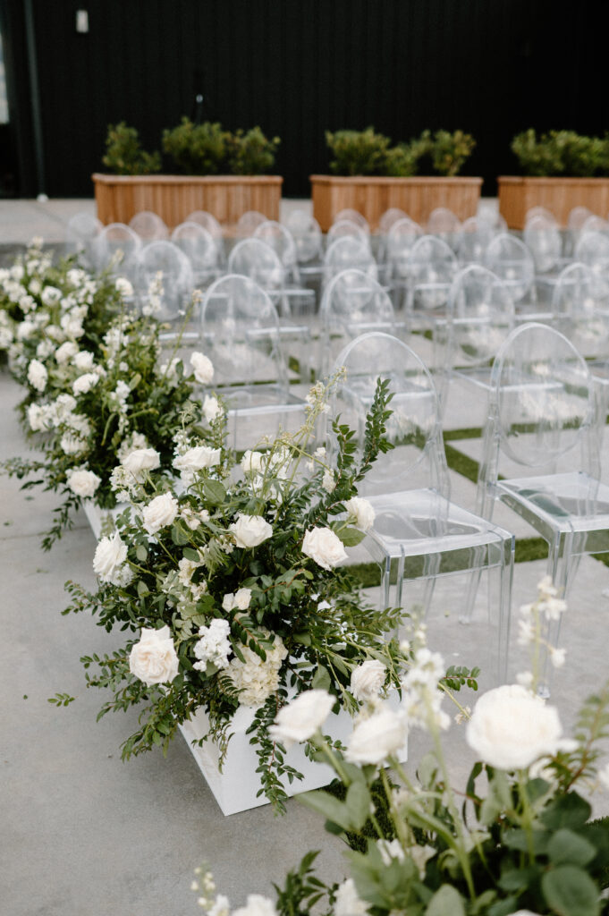Large floral arrangements made with greenery and white roses placed on white boxes next to the clear acrylic chairs going down the aisle for wedding ceremony