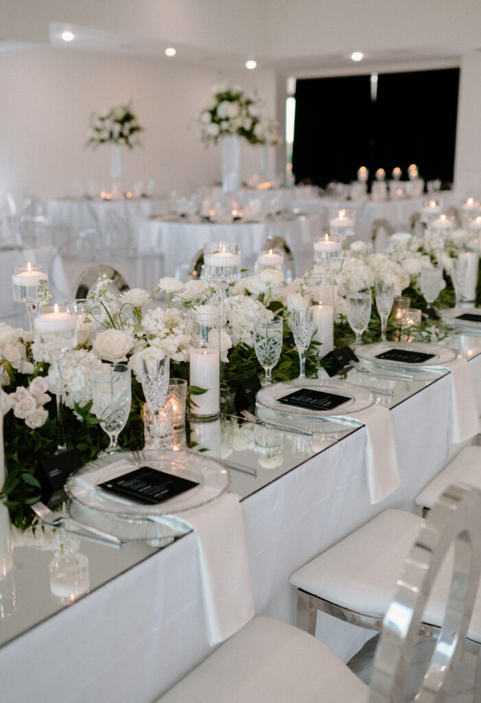 Wedding reception guest tables with a floral runner made of greenery and white flowers mixed with assorted candles 
