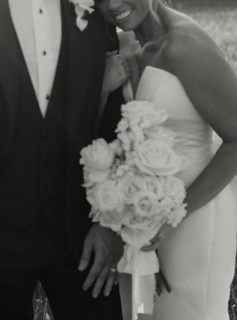 Black and white photo of bride and groom posing with the bride's long stemmed white rose bouquet 