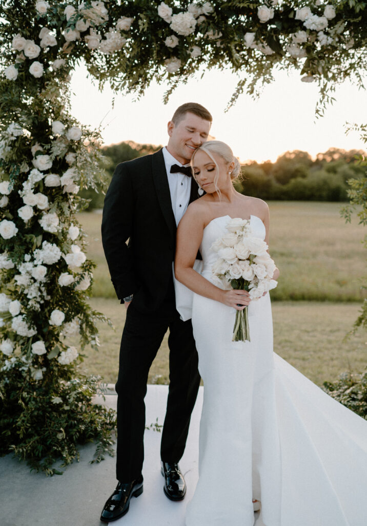 Bride and groom posing with her bouquet made of long stemmed white roses standing in front of the ceremony alter with a large square floral arch made with white roses and greenery