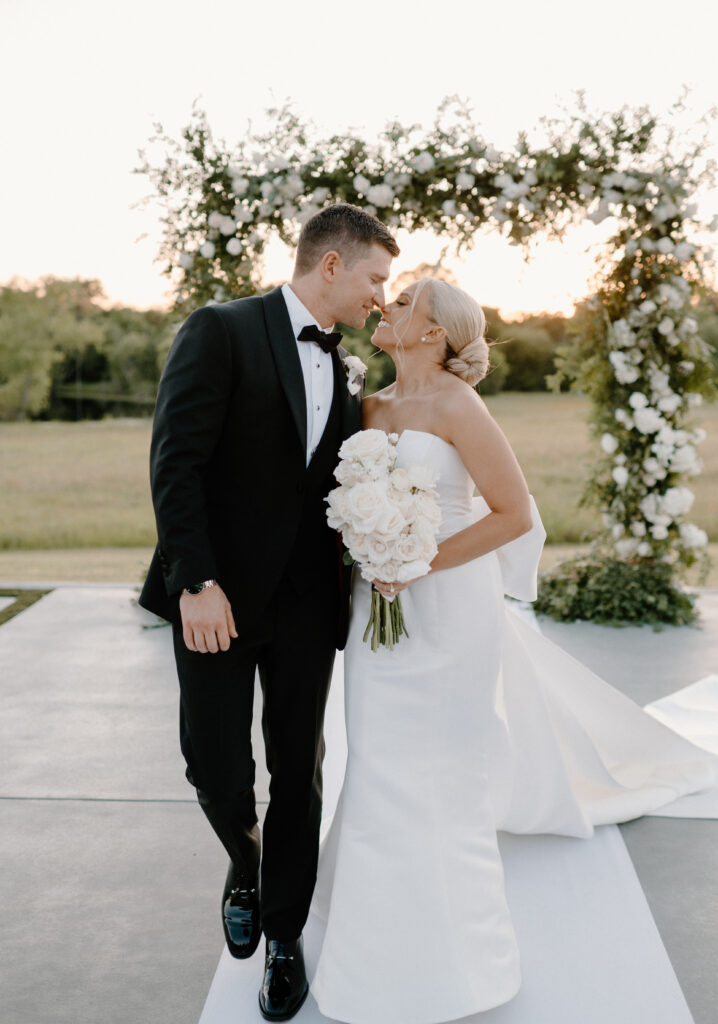 Bride and groom posing with her bouquet made of long stemmed white roses standing in front of the ceremony alter with a large square floral arch made with white roses and greenery