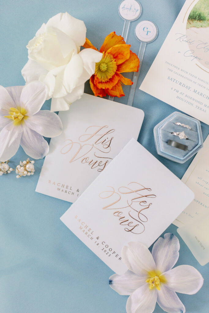 A colorful flat lay with vow books placed on a dusty blue colored back drop with the couples wedding rings and accenting flowers placed around it