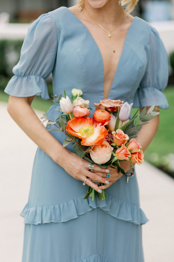 A bridesmaid in a dusty blue dress holding her brightly colored bouquet that includes poppies