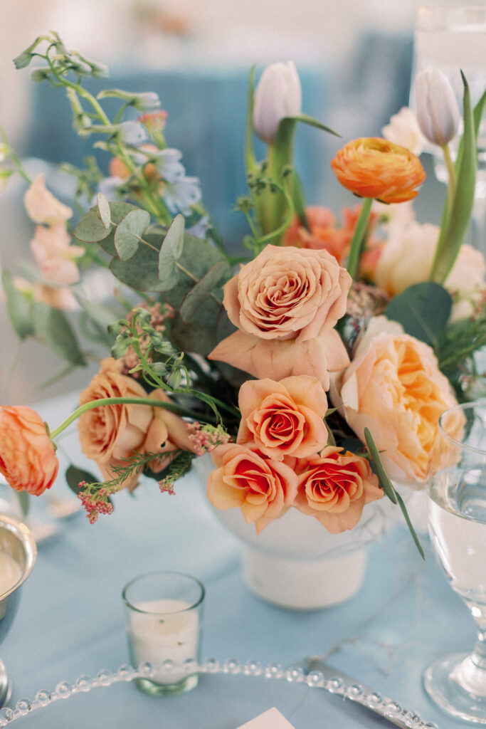 A colorful floral arrangement made with brightly colored flowers in a white compote bowl placed on a dusty blue linen with votive candles for a spring wedding reception