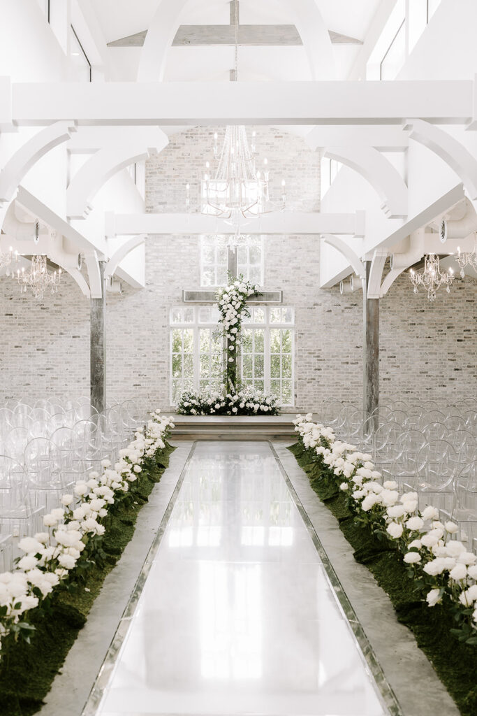An aisle way created of tall white roses leading to a cross of lush florals for a bride and groom to say I do.