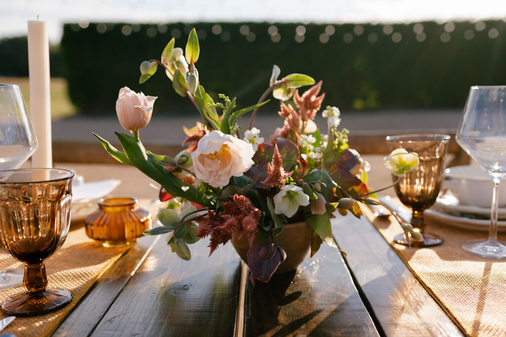 A floral compote made in a wooden bowl with fall colored blooms including roses, celosia, and hellebore used as centerpieces for an elegant fall themed wedding