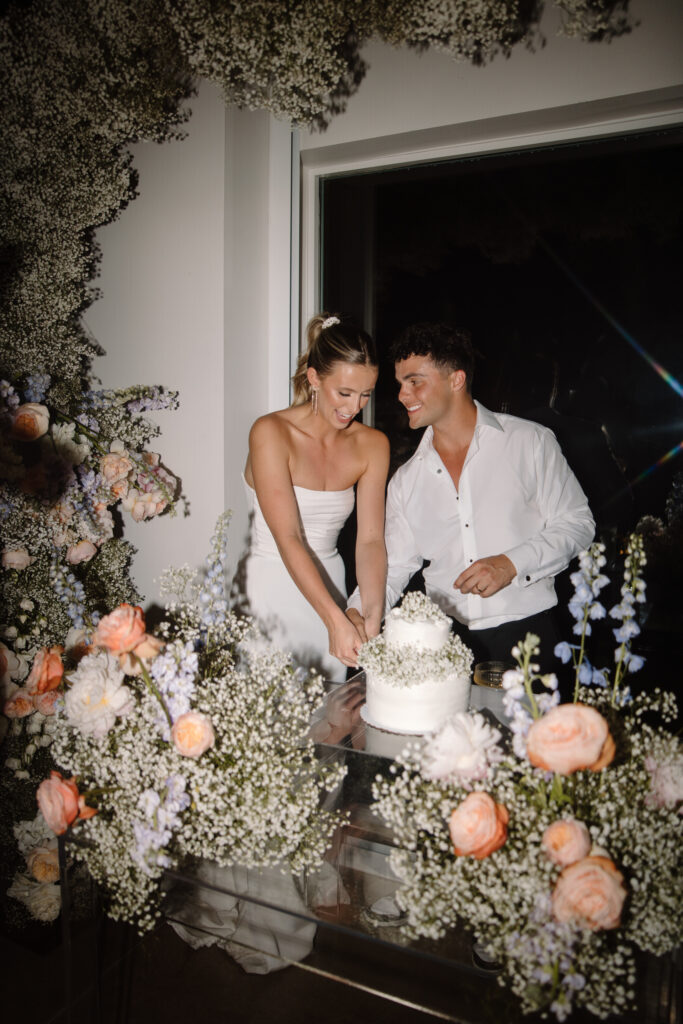 Bride and groom cutting wedding cake under a floral arch made of baby's breath and pastel colored flowers with smaller floral arrangements in front of them