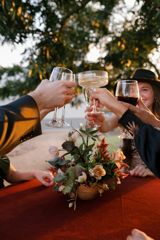 Wedding guest clinking their glasses together to cheers over a floral compote made in a wooden bowl with fall colored blooms including roses, celosia, and hellebore 