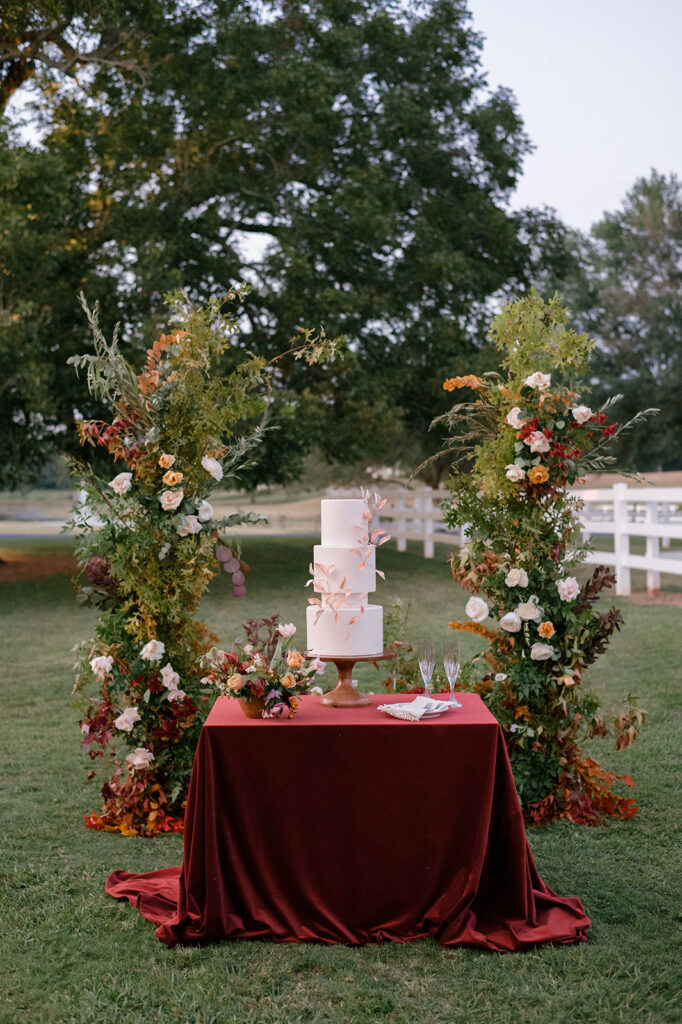 An elegant wedding cake table set up with a repurposed wedding arch behind it made with fall inspired florals