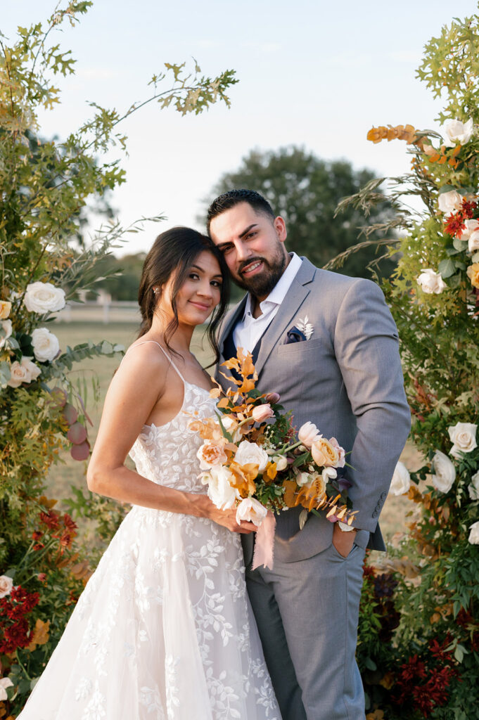 Bride holding her bouquet posing with groom in front of wedding ceremony arch made with fall colored florals