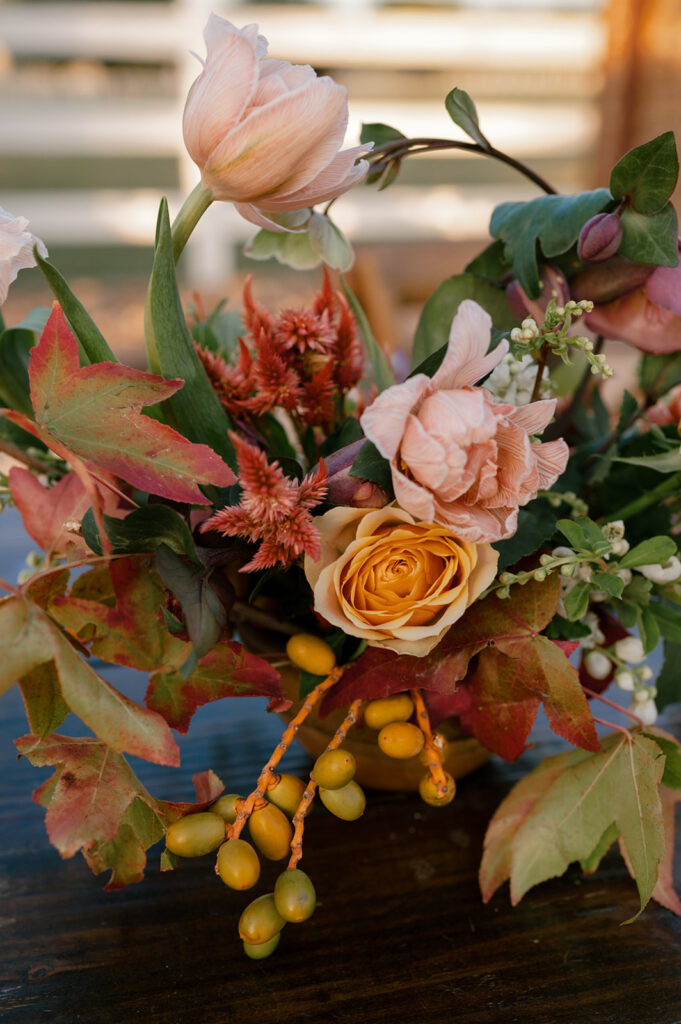 A floral compote made in a wooden bowl with fall colored blooms including roses, celosia, and hellebore used as centerpieces for an elegant fall themed wedding