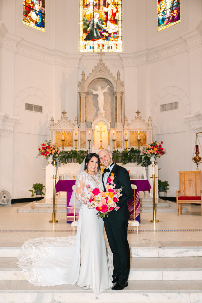 Bride and groom posing with the colorful bridal bouquet made with pink and orange flowers in front of ceremony alter