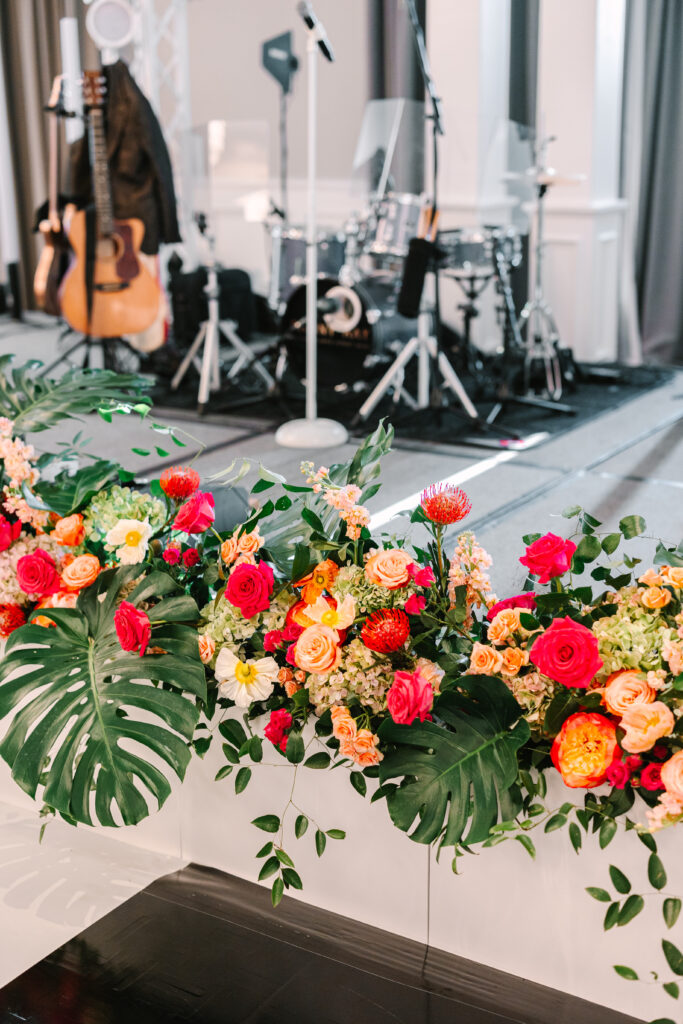 White facades fill with brightly colored orange and pink florals and tropical leaves placed in front of the wedding band stage