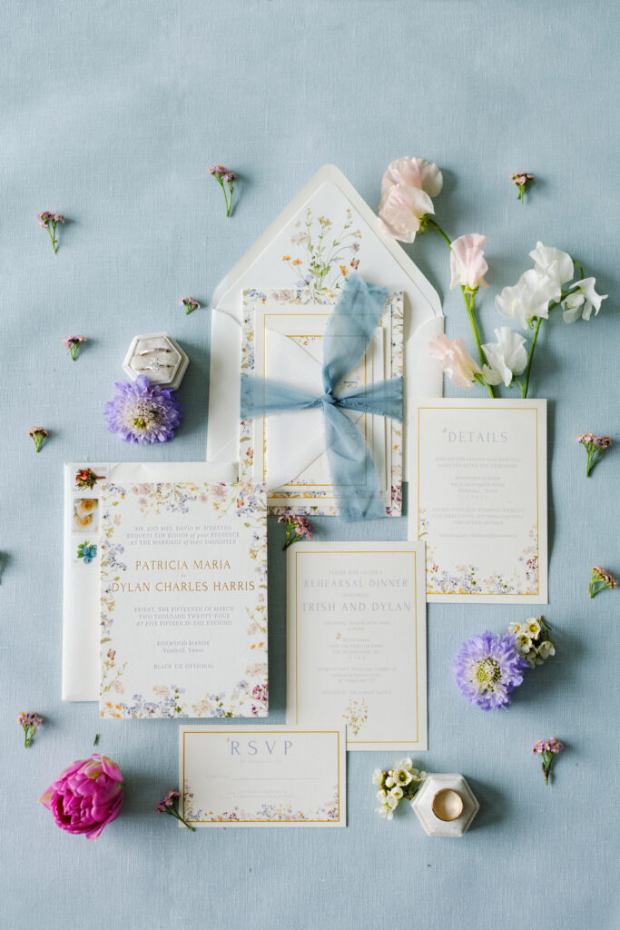 Spring wedding invitation suite with pastel colored blooms placed around, all sitting on light blue flat lay backdrop 