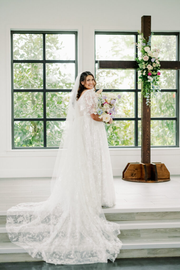 Bride posing with her whimsical pastel colored bouquet and standing in front of the ceremony alter cross with a matching floral arrangement installed 