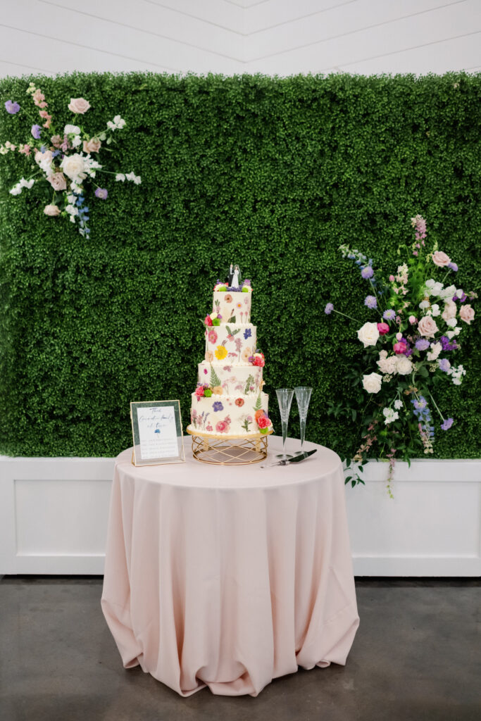 White wedding cake with colorful spring blooms placed on cake sitting in front of a boxwood wall with two accenting floral arrangements placed on wall to frame the cake 