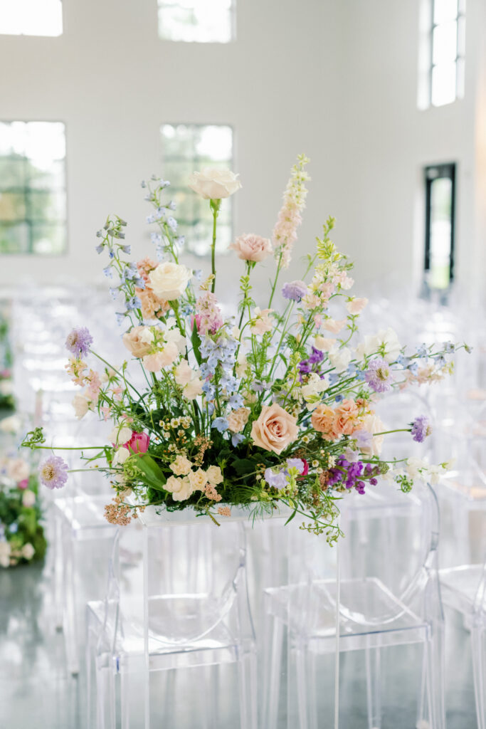 Large textural pastel colored floral arrangement placed on a clear acrylic pedestal at the back of the ceremony aisle 