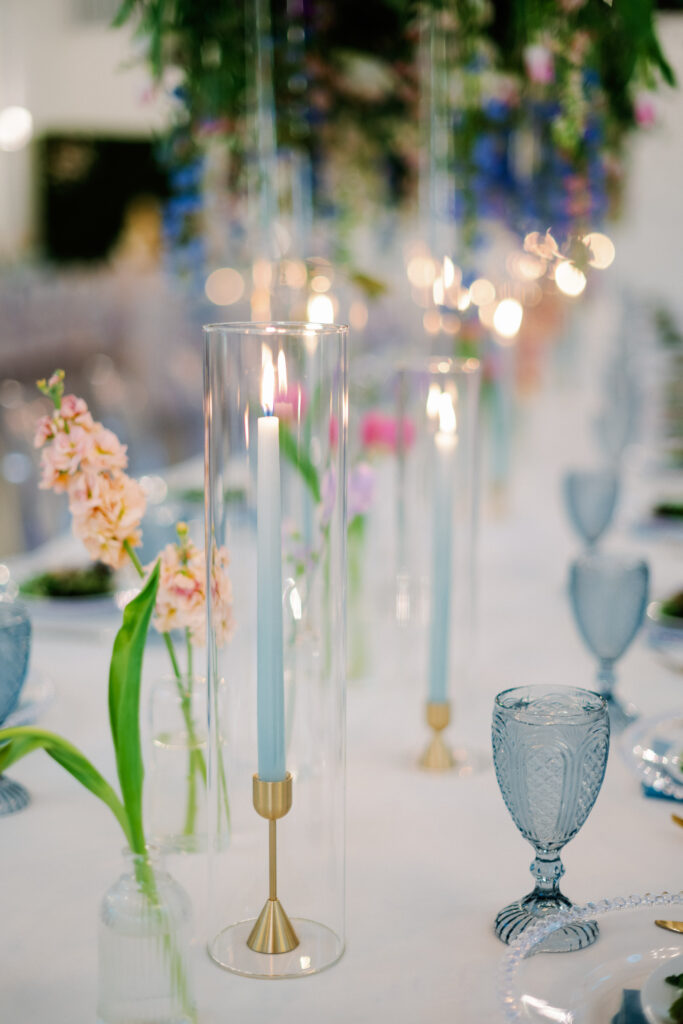 Blue taper candles with gold holders mixed with glass bud vases with pastel colored blooms placed on wedding reception table